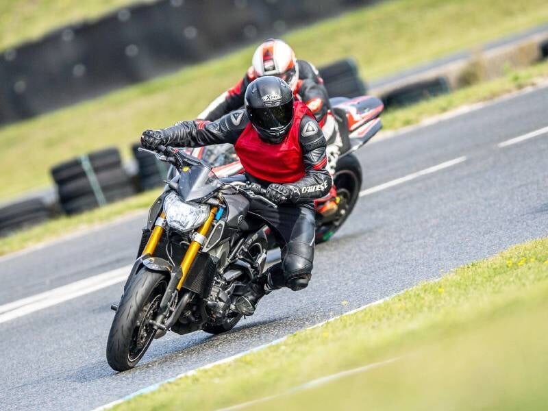 Deux motards en tenue de protection rouge et noire pilotent leurs motos sportives sur un circuit goudronné en virage.