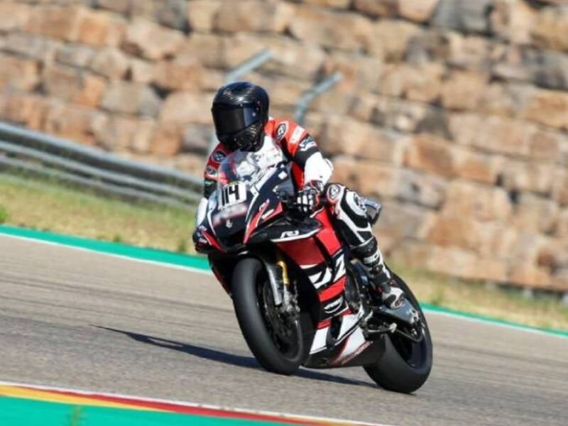 Un pilote en combinaison complète et casque noir roule à grande vitesse sur une moto sportive rouge et noire sur un circuit asphalté.