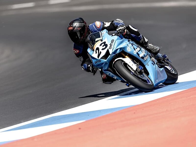 Pilote penché sur une moto bleue claire numérotée 23, attaquant un virage sur une piste avec des bordures bleues et rouges.