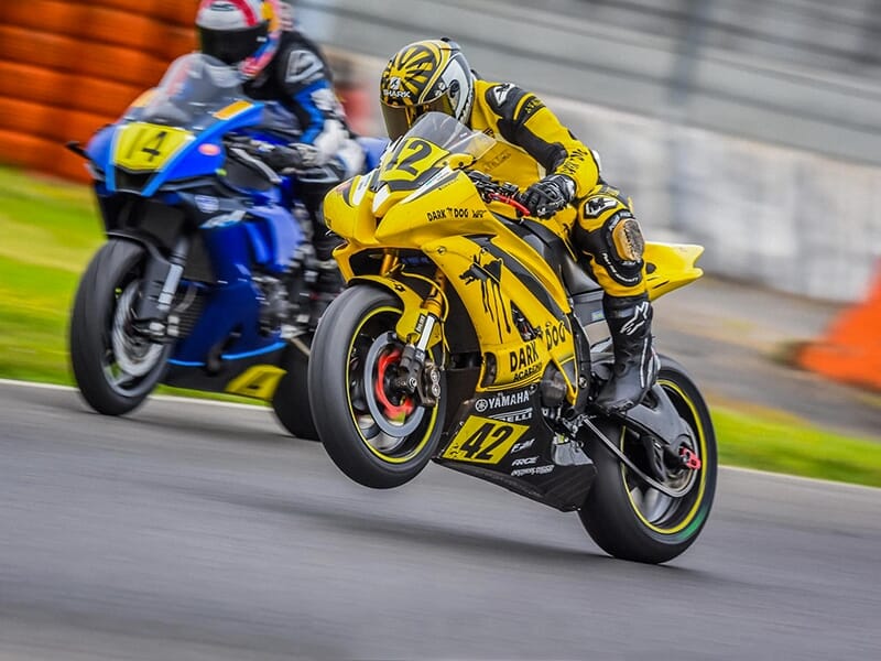 Moto jaune numérotée 42 avec un pilote effectuant un wheeling, suivi d’un autre pilote sur une moto bleue numérotée 14.