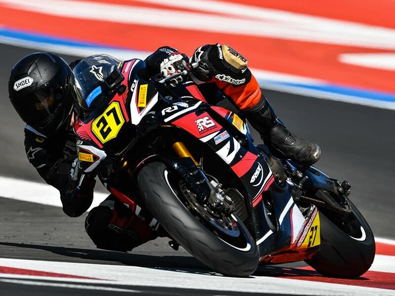 Pilote penché au guidon d’une moto rouge et noire numérotée 27, négociant un virage sur une piste rouge et blanche.