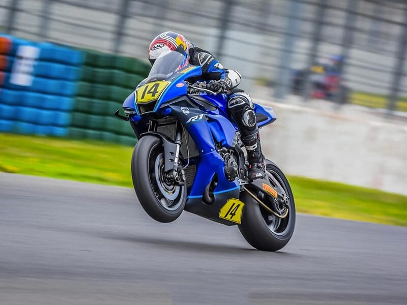 Un pilote sur une Yamaha R1 bleue numérotée 14, en plein wheeling sur un circuit, avec une bordure verte et des pneus empilés en arrière-plan.