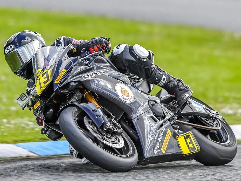 Pilote incliné sur une moto noire numérotée 13, effectuant un virage serré sur une piste bordée d’herbe.