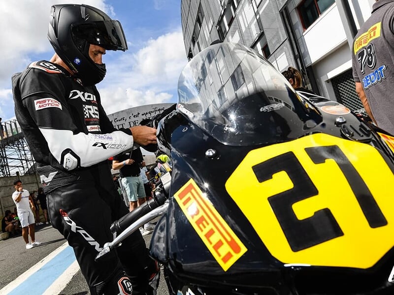Un pilote en combinaison noire et casque, ajustant les réglages de sa moto numéro 21, garée dans la zone des stands, entouré de spectateurs et d'autres participants.