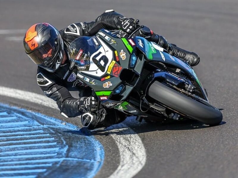 Un pilote en combinaison noire, au guidon d’une moto verte et noire numérotée 16, prenant un virage avec une inclinaison extrême, frôlant le sol.