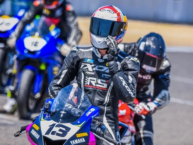 Un pilote en combinaison et casque de moto au premier plan, debout sur une Yamaha numéro 13, avec d'autres motos alignées en arrière-plan sur un circuit.