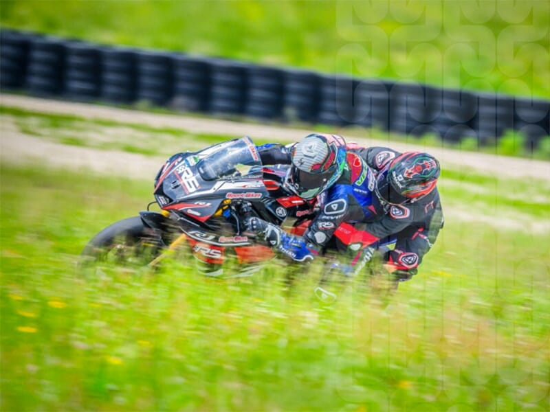 Deux pilotes en combinaison de course inclinés sur une moto, prenant un virage serré à pleine vitesse, avec un décor de nature verdoyante en arrière-plan.