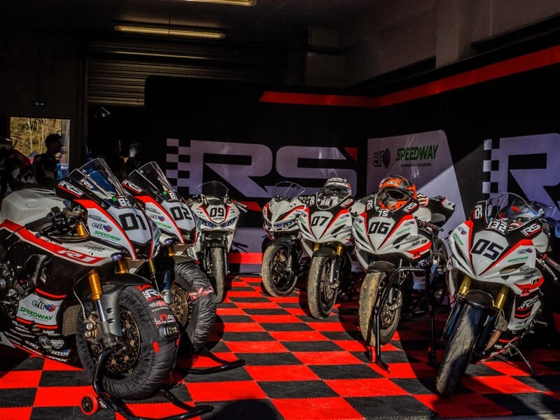Cinq motos de course blanches alignées dans un garage avec un sol à damier rouge et noir, prêtes à rouler.