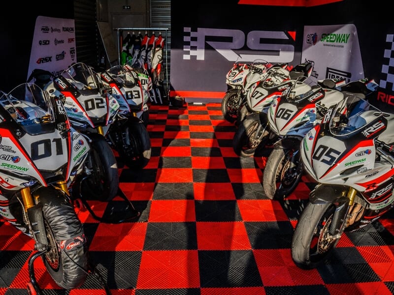 Six motos de course blanches alignées dans un garage avec un sol à damier rouge et noir, ambiance sportive.