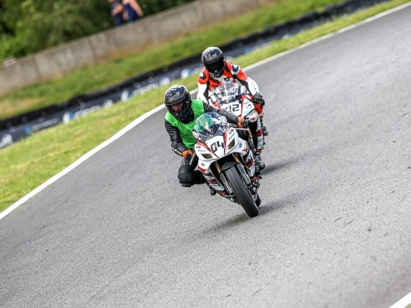 Deux motards en tenue de course sur leurs motos blanches, roulant côte à côte sur un circuit asphalté.