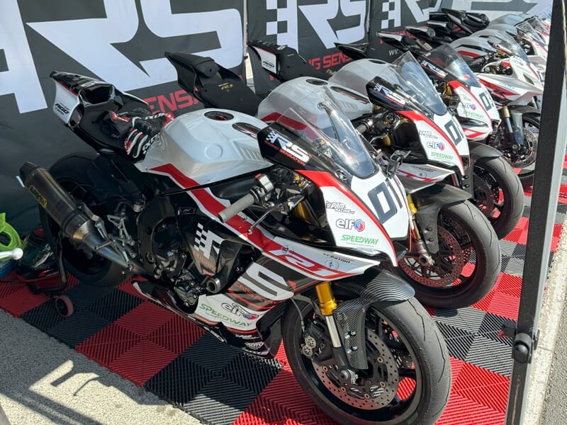 Plusieurs motos de course blanches alignées dans un garage avec un sol à damier rouge et noir.