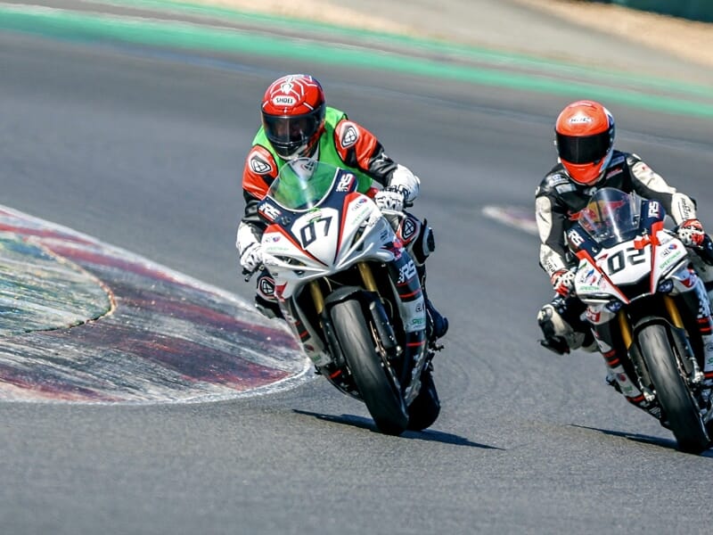 Deux pilotes en combinaison rouge et blanche sur leurs motos blanches, prenant un virage sur circuit.