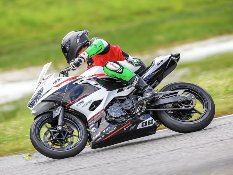 Motard en combinaison verte et noire sur une moto blanche, inclinée dans un virage sur piste de course.