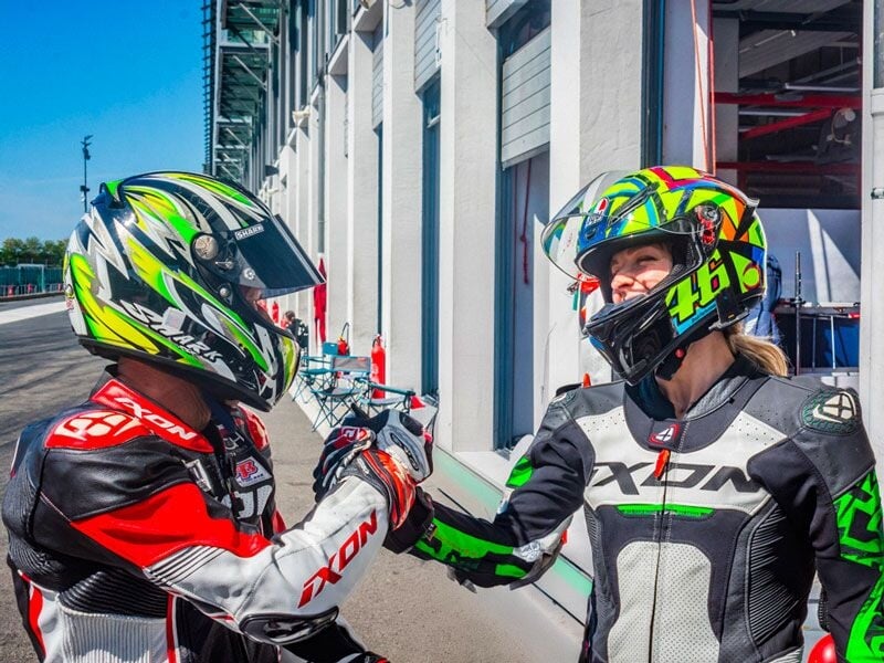 Deux motards en tenue complète et casques colorés se serrent la main devant un bâtiment blanc au circuit de Bresse.