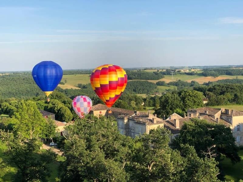 Trois montgolfières colorées flottent au-dessus d'un village entouré de verdure sous un ciel bleu clair.