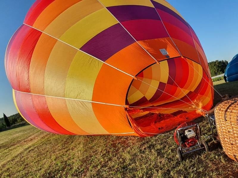 Montgolfière aux rayures rouges, jaunes et orange en train de se gonfler sur un champ herbeux au coucher du soleil.