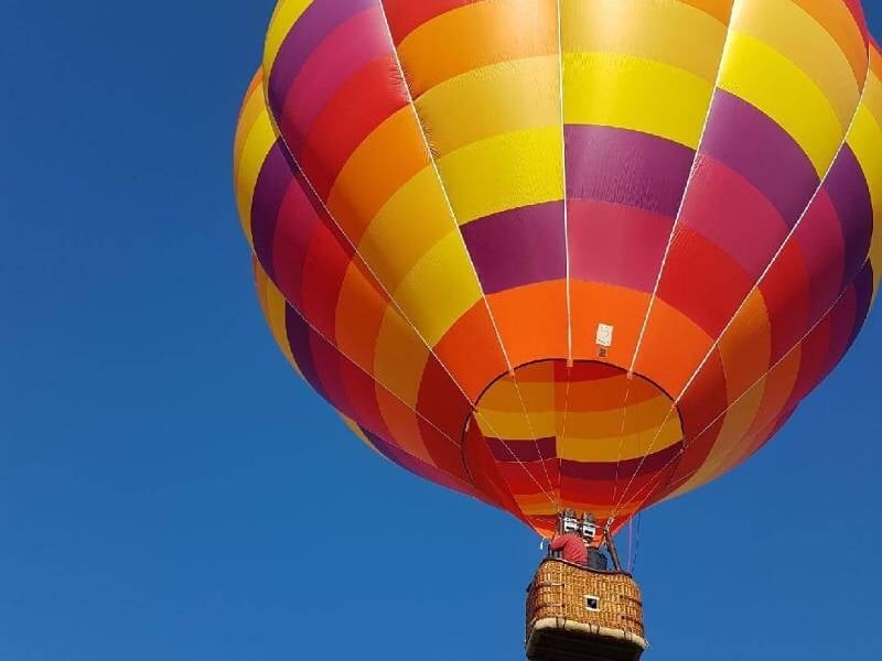 Montgolfière rouge, jaune et orange en vol avec une nacelle suspendue, sur fond de ciel bleu sans nuages.