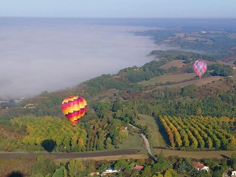 Deux montgolfières colorées survolent une vallée verdoyante avec des collines et une légère brume matinale.