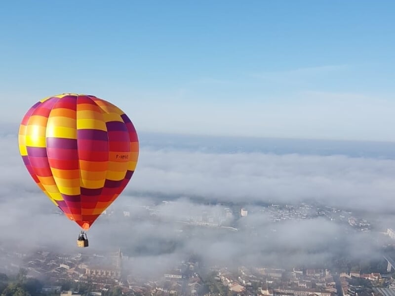 Montgolfière rouge, jaune et orange flottant au-dessus d'une ville partiellement couverte de brouillard sous un ciel clair.
