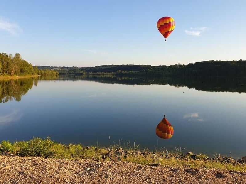 Montgolfière rouge, jaune et orange se reflète dans l'eau calme d'un lac entouré d'arbres verts au bord du rivage.