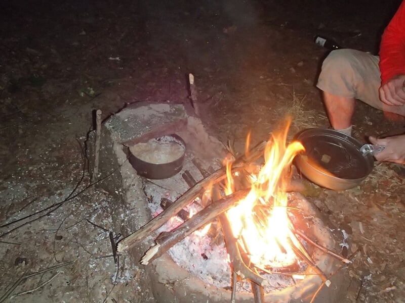 Feu de camp allumé dans un foyer en pierre avec une casserole posée dessus, personne à côté en tenue de survie.