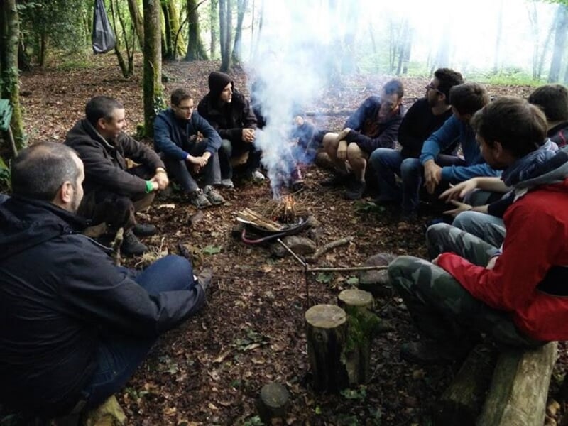 Groupe de personnes assises en cercle autour d'un feu de camp fumant dans une forêt dense, ambiance conviviale.
