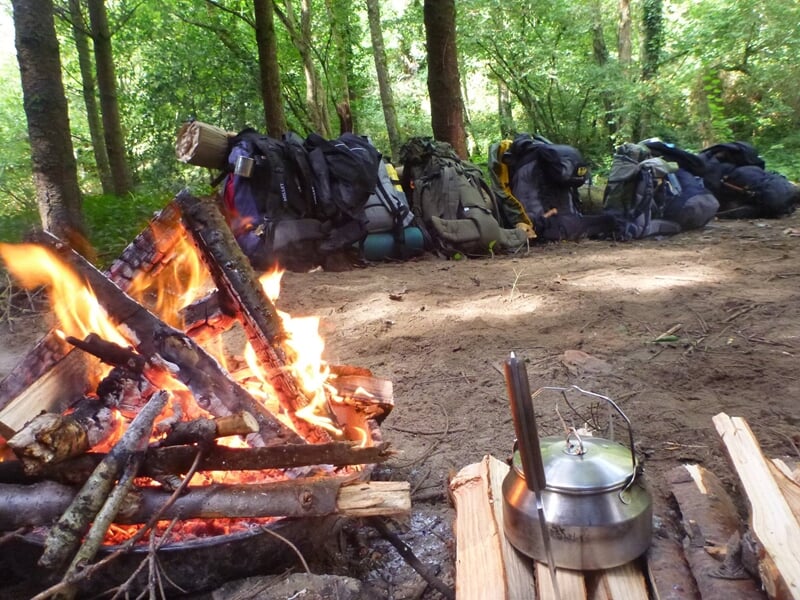 Feu de camp avec des bûches enflammées, plusieurs sacs à dos posés au sol dans une clairière boisée.