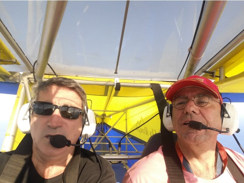 Deux hommes dans un ULM à cockpit ouvert, équipés de casques audio, l'un portant une casquette rouge, sous une structure jaune et bleue