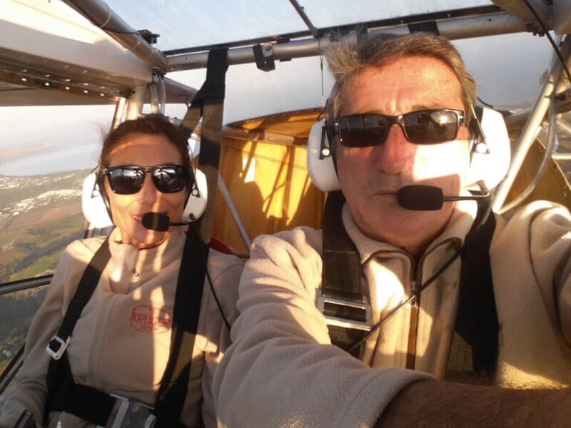 Deux personnes dans un cockpit d'ULM, portant des casques avec micro, lunettes de soleil et vêtements de vol beige, lumière douce du matin ou du soir