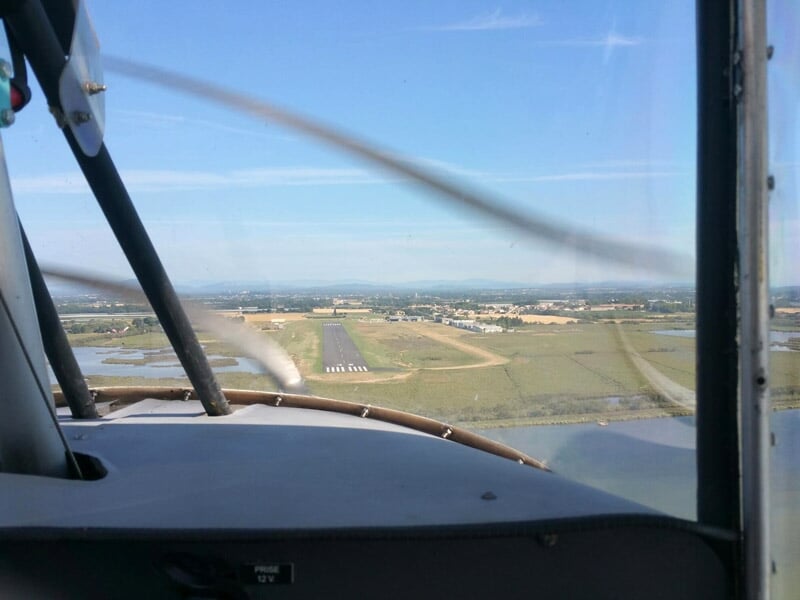 Approche d'une piste d'atterrissage vue depuis l'intérieur d'un cockpit, avec paysage rural environnant, marais et collines à l'horizon