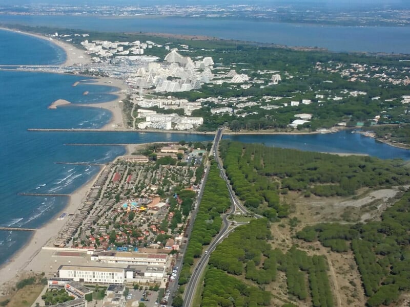 Vue aérienne d'une station balnéaire avec plages, immeubles en forme de pyramide, et espaces boisés entourant un cours d'eau