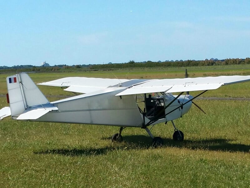 Avion léger blanc stationné sur une piste en herbe, sous un ciel bleu dégagé, avec un drapeau français sur la dérive arrière