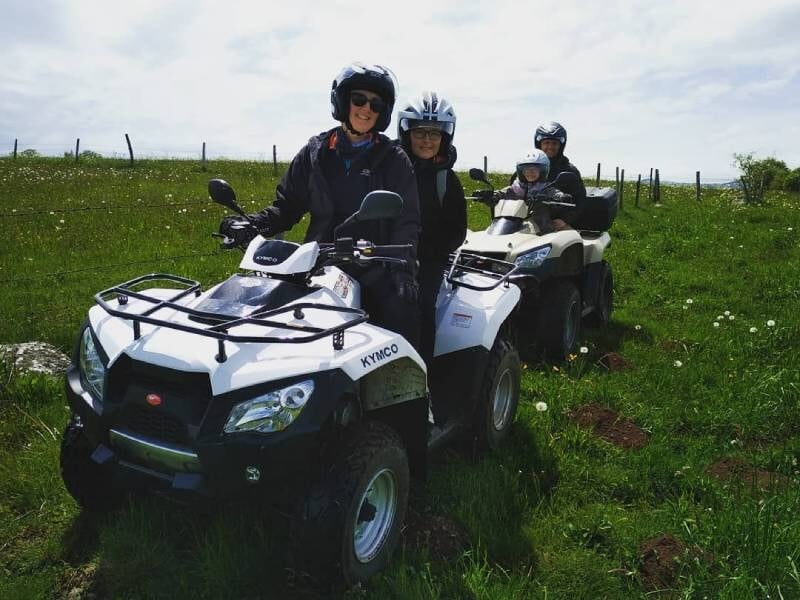 Deux personnes en tenue de protection sur un quad blanc dans un champ vert sous un ciel nuageux.