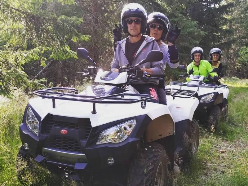 Quatre personnes portant des casques sur des quads blancs traversant une forêt dense et ensoleillée.