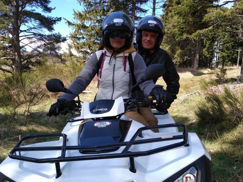 Couple portant des casques assis sur un quad blanc dans une forêt avec des arbres et de la lumière.