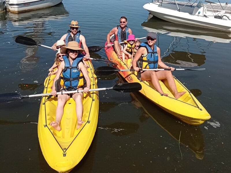 Quatre personnes en gilets de sauvetage pagayant dans deux kayaks jaunes sur une rivière calme près d'un barrage.