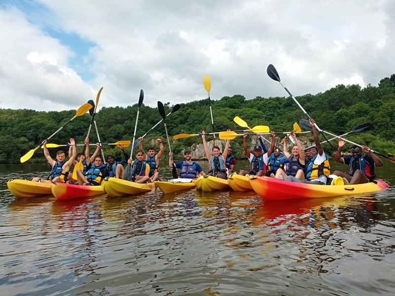 Groupe de personnes en kayak jaune sur une rivière, levant leurs pagaies en signe de joie sous un ciel partiellement nuageux.