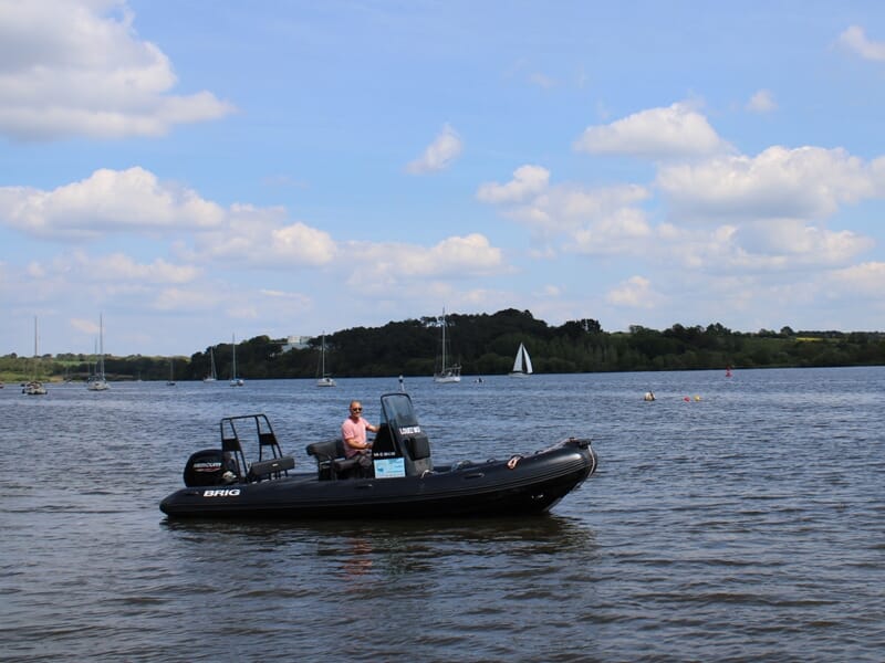 Un homme sur un bateau pneumatique noir navigue sur une rivière calme sous un ciel bleu avec des nuages blancs.