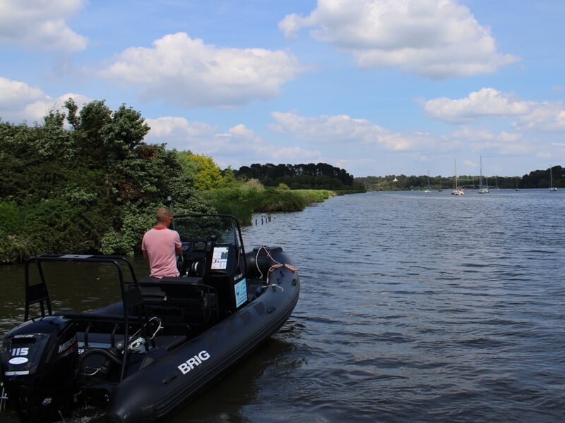 Un homme en t-shirt rose est assis sur un bateau pneumatique noir amarré près de la rive avec des arbres verts.