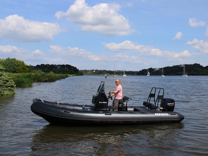 Un homme debout sur un bateau pneumatique noir sur une rivière avec des voiliers et des arbres en arrière-plan.