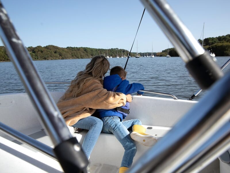 Une femme et un enfant assis dans un bateau sur un lac calme, entourés de nature et de ciel bleu.
