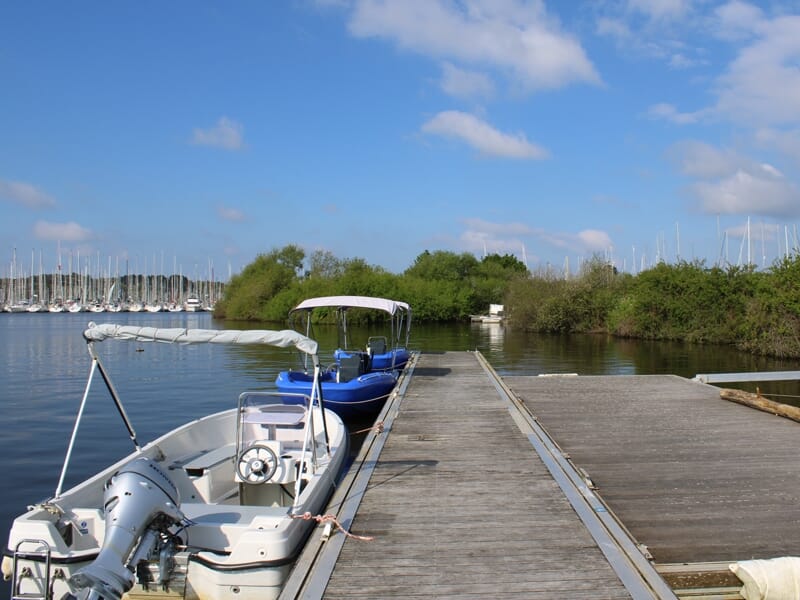 Un ponton en bois avec plusieurs petits bateaux amarrés, sous un ciel bleu avec quelques nuages.