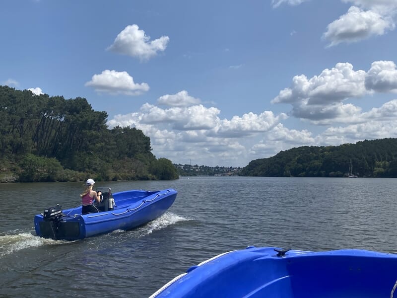 Une personne conduisant un petit bateau bleu sur une rivière sous un ciel partiellement nuageux.