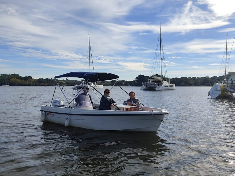 Trois personnes dans un petit bateau blanc sur une rivière avec plusieurs voiliers et un ciel bleu.