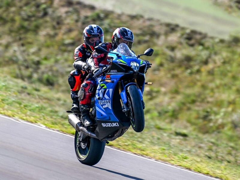 Deux motocyclistes exécutant une figure en tandem sur une moto sportive Suzuki bleue, le passager arrière debout et saluant, sur une route de campagne