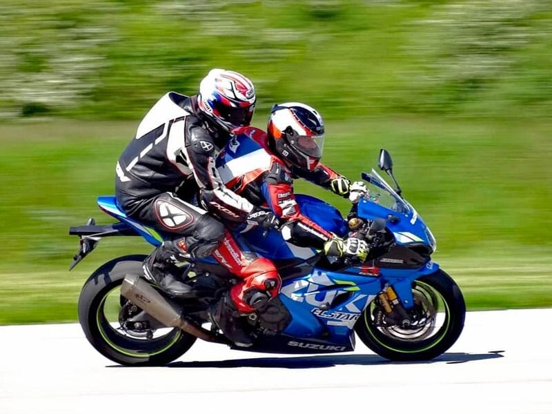 Deux motocyclistes en tenue de course noire, blanche et rouge sur une moto sportive Suzuki bleue, roulant côte à côte sur une route avec de l'herbe verte en arrière-plan