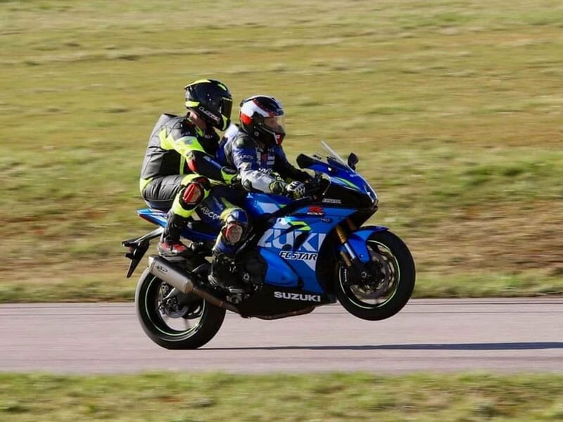 Deux motocyclistes, un en jaune fluo et l'autre en bleu, sur une moto sportive Suzuki, en course sur une piste avec de l'herbe sèche en arrière-plan