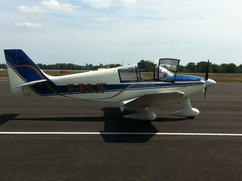 Petit avion blanc et bleu stationné sur une piste d'atterrissage sous un ciel nuageux clair.