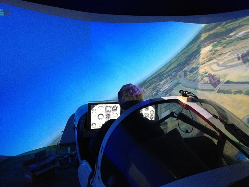 Personne dans un simulateur de vol avec un écran panoramique montrant un paysage aérien.