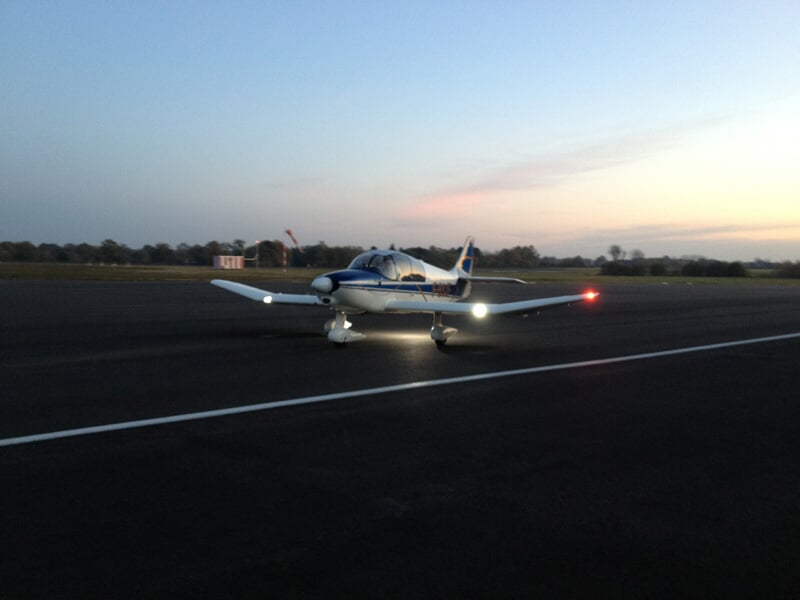 Avion blanc et bleu sur une piste au coucher du soleil avec les feux de navigation allumés.